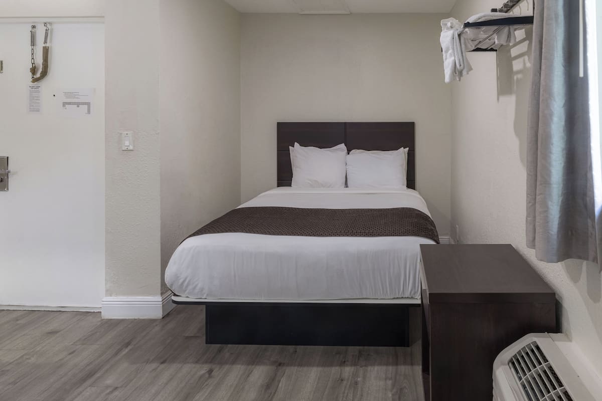 A hotel room with a bed, a bedside table, a wall-mounted heater, and a door with a towel rack.