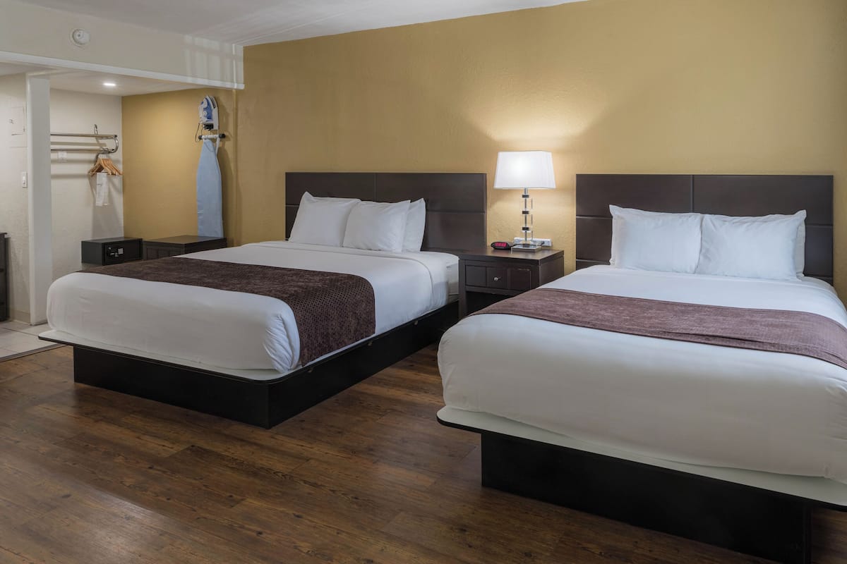 A hotel room with two beds, wooden flooring, a nightstand with a lamp, and a coat rack.