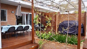 A wooden deck with a table and chairs, a covered barbecue area, and a brick wall with climbing plants. - Eden by the Bay, 98 Sandy Point Rd - Large family home just 300m to Corlette Beach (Corlette)