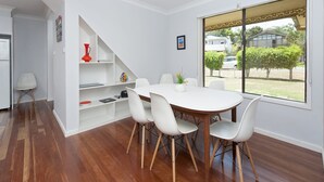 A dining area with a white table and chairs, a wooden floor, and a view of the outside through a large window. - Eden by the Bay Faimly Home near Corlette Beach (Corlette)