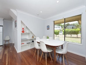 A dining area with a white table and chairs, a wooden floor, and a view of the outside through a large window. - Eden by the Bay Faimly Home near Corlette Beach (Corlette)