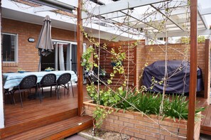 Un espace extérieur abrité, doté d’une terrasse en bois, d’une table avec des chaises et d’une structure pour plantes grimpantes.