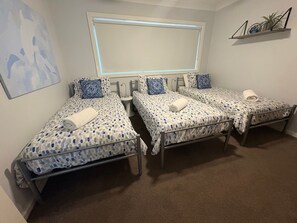 A hotel room with three single beds, each with a patterned bedspread and matching pillows. A large window with blinds is above the beds, and a small shelf with decorative items is on the wall.