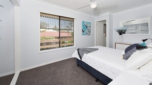 A bedroom with a bed, a ceiling fan, a window with a view, a painting, and a mirror. - Eden by the Bay Faimly Home near Corlette Beach (Corlette)
