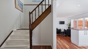 A staircase with wooden balusters leading to a landing, a flat-screen TV mounted on the wall, and a kitchen area with a dining table and chairs. - Eden by the Bay, 98 Sandy Point Rd - Large family home just 300m to Corlette Beach (Corlette)