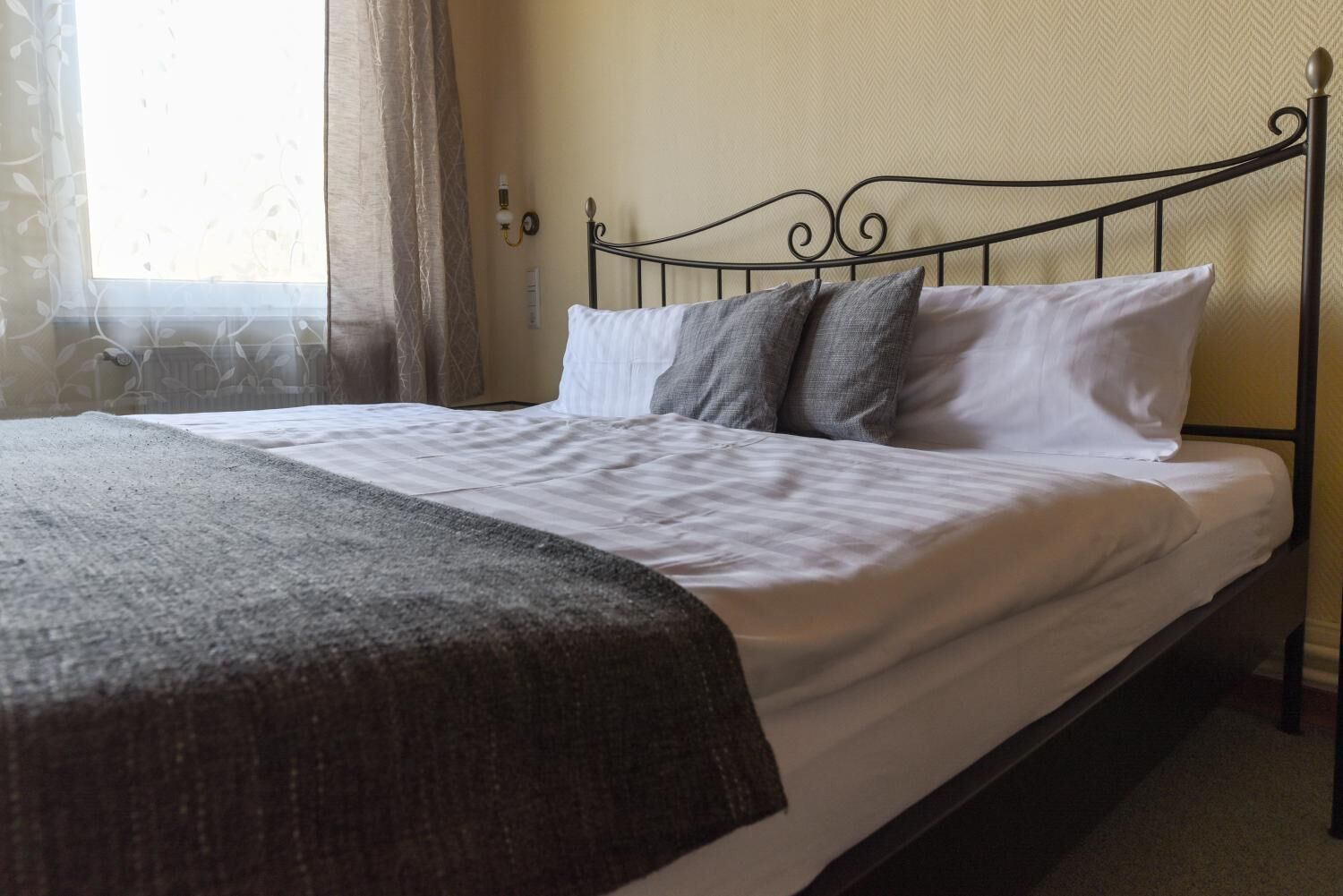 A neatly made bed with white linens and a dark throw blanket, a metal headboard, and a window with sheer curtains.