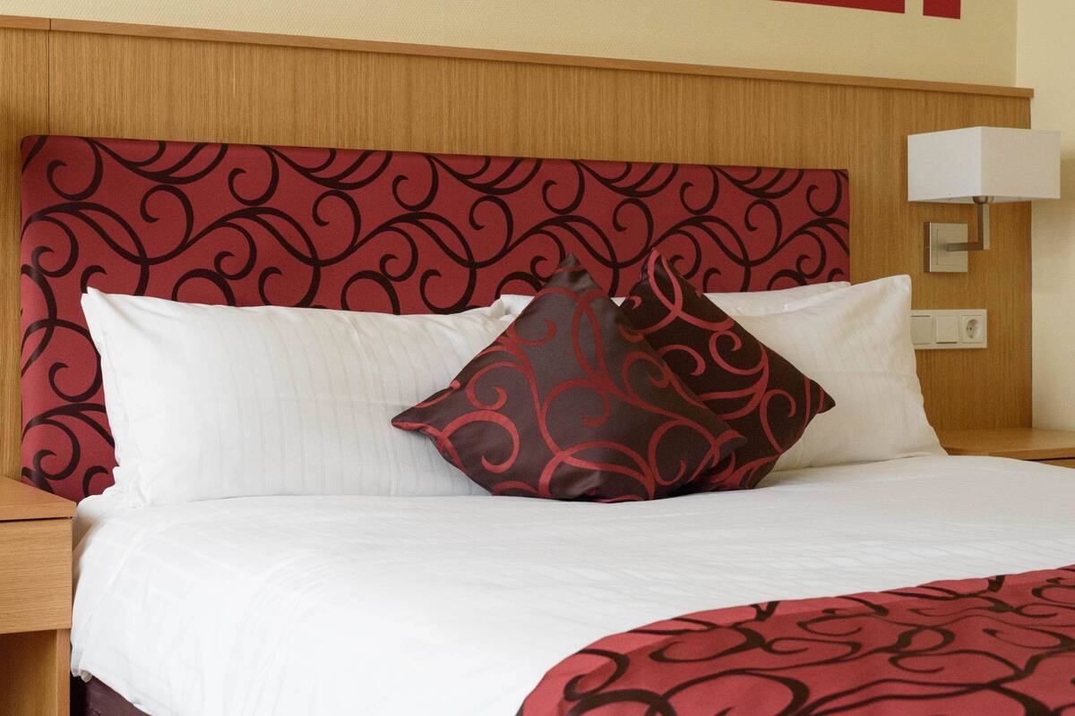 A hotel room with a bed, white bedding, a red patterned headboard, and a bedside lamp.