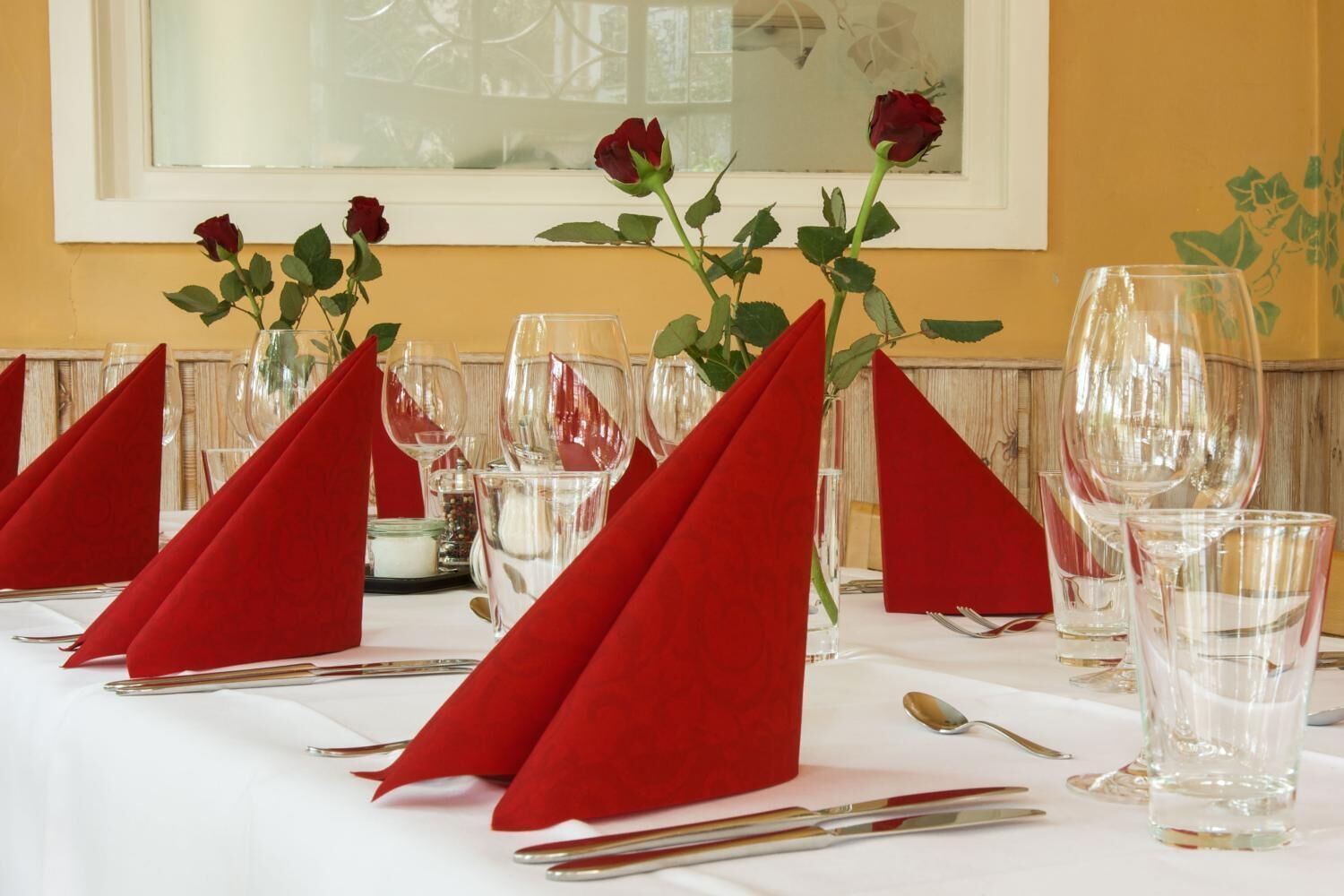 Una mesa de comedor con servilletas rojas, copas de vino transparentes y un jarrón con rosas rojas.