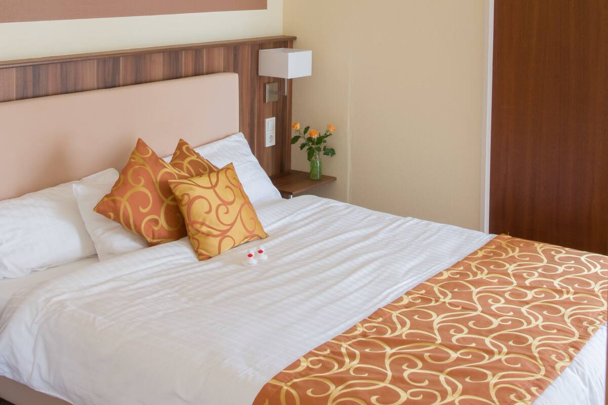 A neatly made bed with white and gold patterned bedding, a wooden headboard, and a bedside table with a vase of flowers.