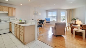 A modern kitchen with wooden cabinets and a central island, a dining area with a TV, and a living room with a sofa and lamp. - Amadeus 2 Top location, water font, steps to beach, restaurants and shopping! (Treasure Island)