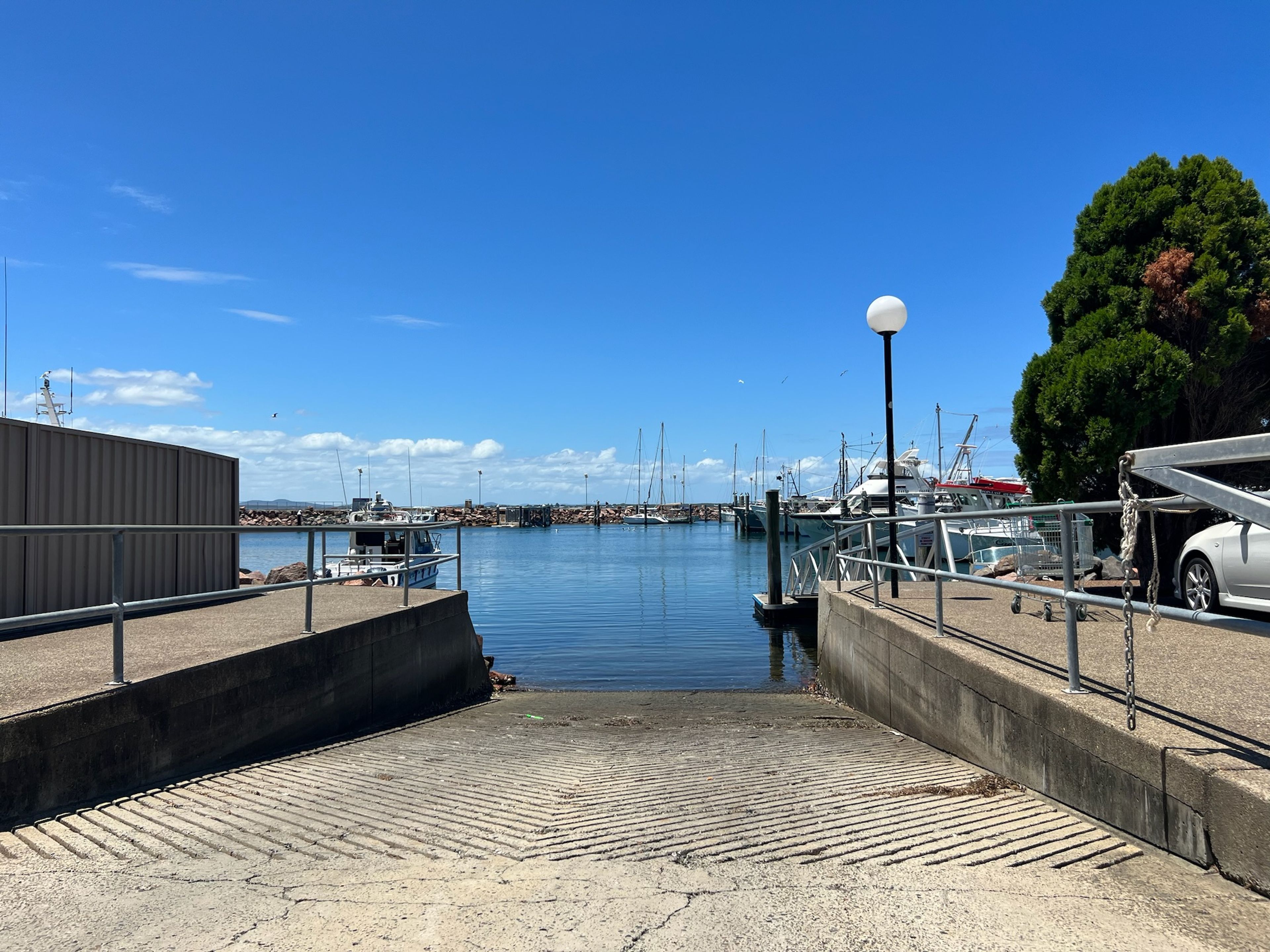 Un port de plaisance avec des bateaux amarrés, une rampe en béton menant à l’eau et un ciel bleu dégagé.
