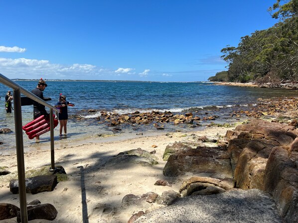 A beach with clear water, rocks, and a metal railing.