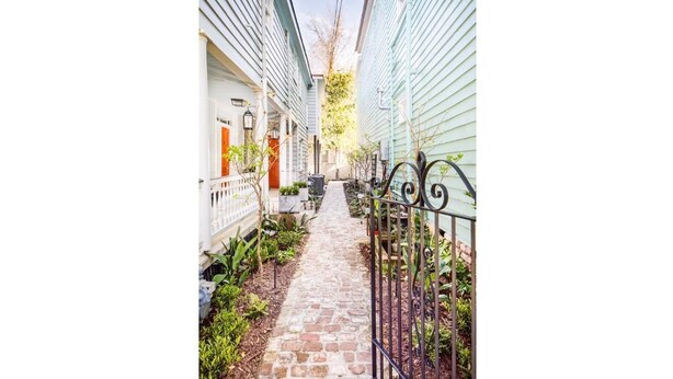 A narrow brick pathway flanked by buildings with white siding and a black wrought iron fence.