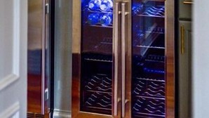 Una cocina con un estante de vidrio que sostiene copas de vino y una botella. Debajo del estante, hay dos neveras para vino.