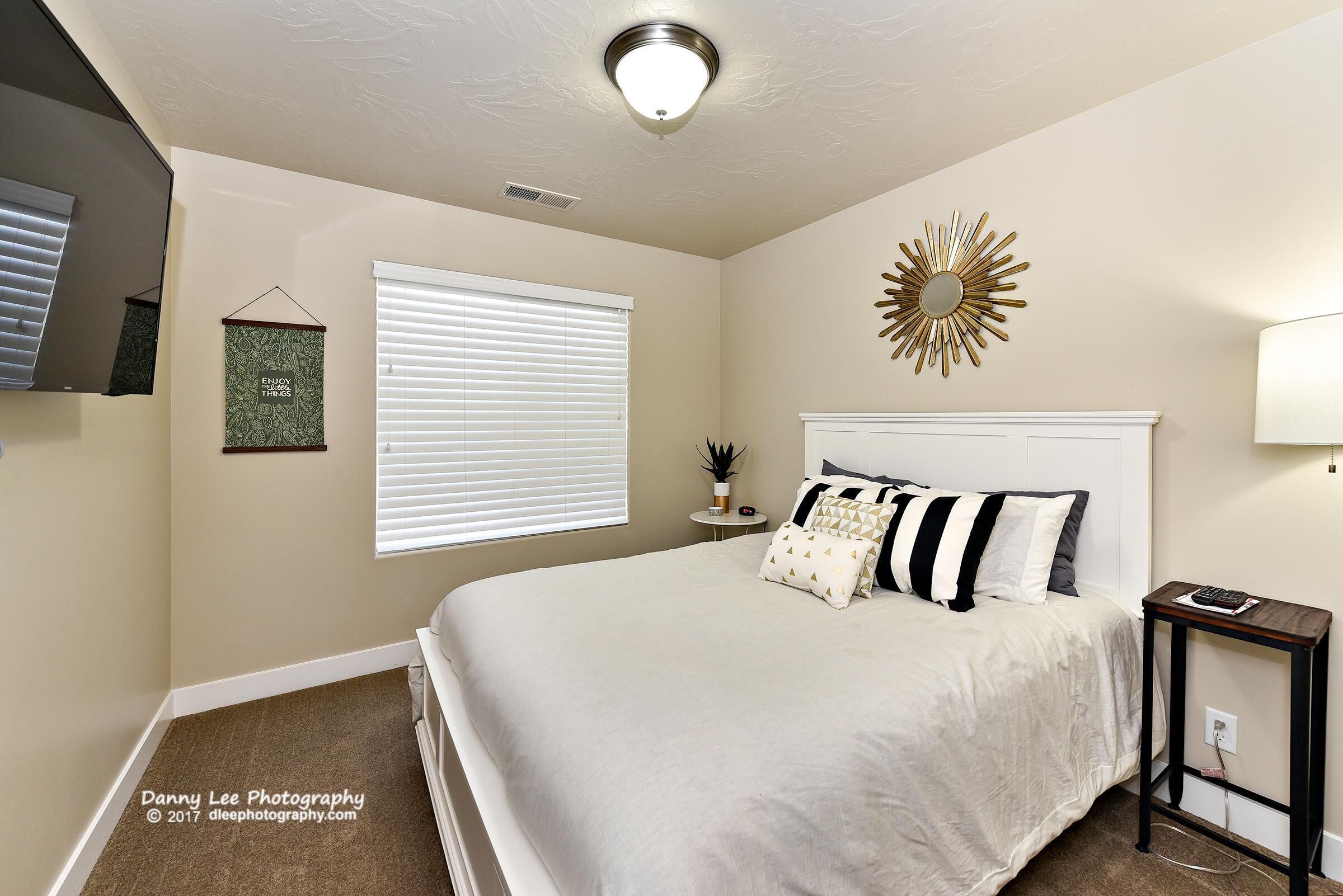 A bedroom with a large bed, a bedside table, a wall-mounted mirror, a window with blinds, and a ceiling light fixture.