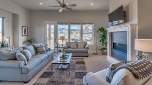 A living room with a sofa, coffee table, and fireplace, with a view of mountains through large windows. - Quail Corner (St. George)