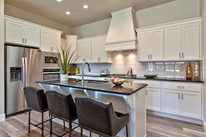 A modern kitchen with white cabinetry, a stainless steel refrigerator, a built-in oven, a large island with bar stools, and a potted plant. - Back Nine Villa (St. George)