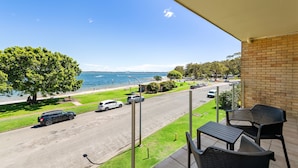 A balcony with outdoor furniture overlooking a street and a beach. - Little Hut Waterfront Apartment in Nelson Bay (Nelson Bay)