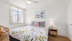 Un dormitorio con una cama, dos mesitas de noche con lámparas, una silla, una ventana con persianas y un ventilador de techo.