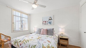 A bedroom with a bed, a nightstand, a chair, a window with blinds, a ceiling fan, and a wall-mounted picture. - Little Hut Waterfront Apartment in Nelson Bay (Nelson Bay)