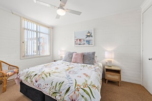 Une chambre à coucher comprenant un lit, une table de chevet, une chaise, une fenêtre avec des stores, un ventilateur de plafond et un tableau accroché au mur.