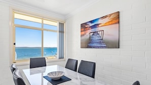 A dining area with a table, chairs, and a view of the sea through a large window. - Little Hut Waterfront Apartment in Nelson Bay (Nelson Bay)