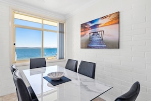 Un espace repas comprenant une table, des chaises et une vue sur la mer, visible à travers une grande fenêtre.