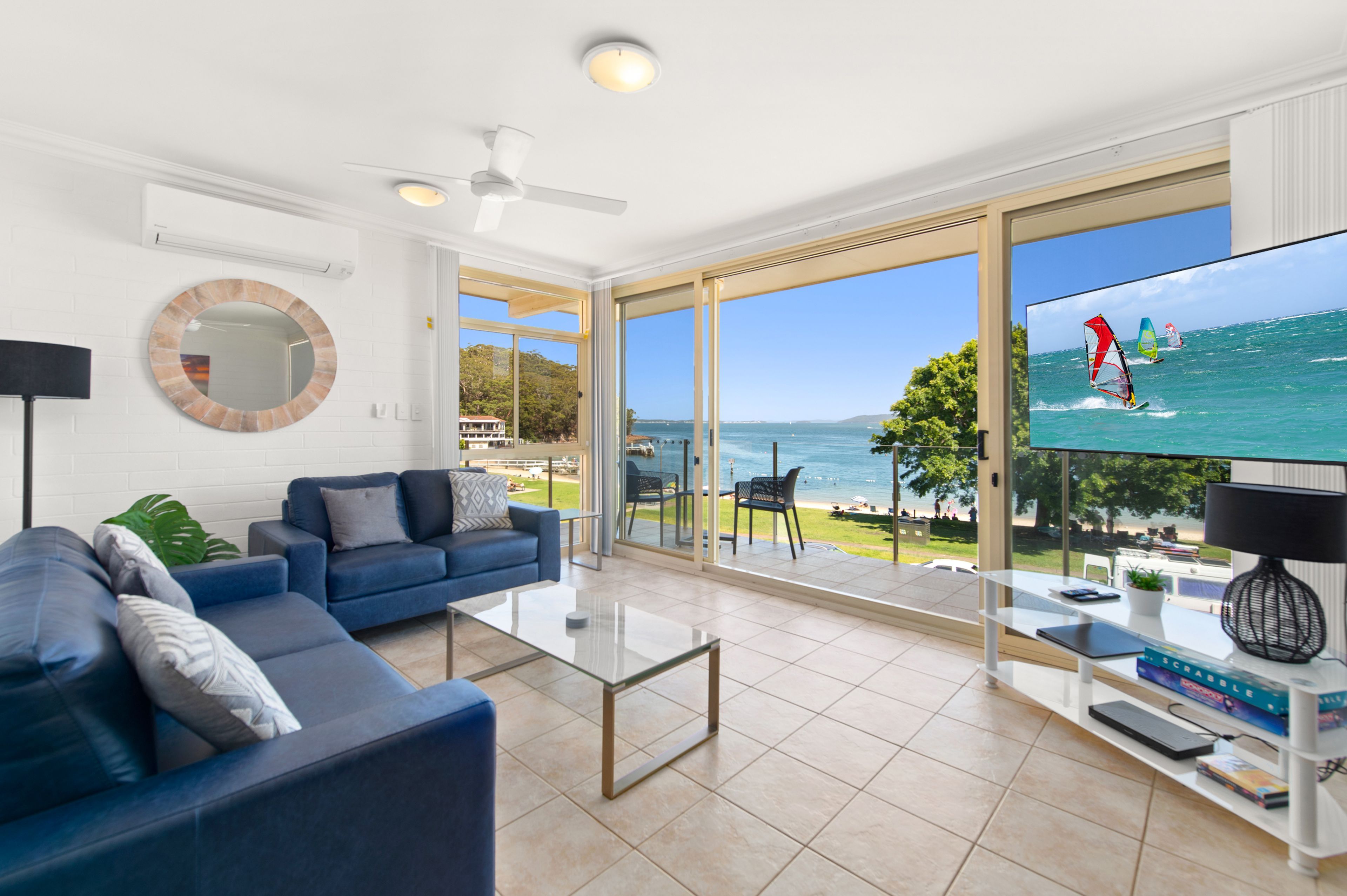 A modern living room with a large TV displaying a beach scene, a sofa, a coffee table, and a view of the sea through large windows.