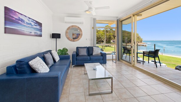 A modern living room with a blue sofa, a coffee table, and a view of the sea through large glass doors. - Little Hut Waterfront Apartment in Nelson Bay (Nelson Bay)