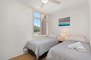 A bedroom with two beds, a window with a view of a building and trees, a painting on the wall, and a ceiling fan.
