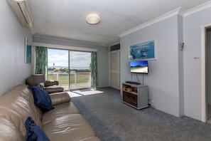 Una sala de estar con un sofá color beige, un televisor sobre un mueble bajo y vistas al exterior a través de un ventanal corredero.
