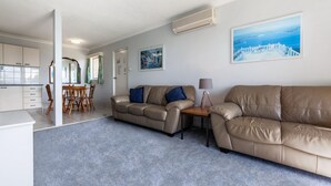 A modern living room with two beige sofas, a wooden dining table, and a kitchen area in the background. - Castaways, 1/17 Shoal Bay Rd - Ground floor, 2-bedroom apartment right across from Shoal Bay Beach (Shoal Bay)