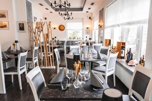 A dining area with multiple tables and chairs, a chandelier, and a view of the outdoors through large windows. - Hotel Restaurant Cala Luna (Marburg)