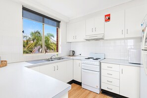 A modern kitchen with white cabinets, a stove, and a window with a view of palm trees. - Seaspray 3 close to Shoal Bay Beach (Nelson Bay)