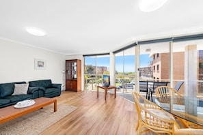 A modern living room with a sofa, coffee table, and a glass dining table with chairs. There is a wooden cabinet and a large window offering a view of the outside. - Seaspray 3 close to Shoal Bay Beach (Nelson Bay)