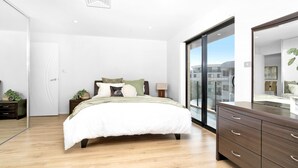 A modern bedroom with a large bed, a dresser, a mirror, and a view of the city through a glass door. - Dolphin Cove 13 Luxury Apartment in Nelson Bay (Nelson Bay)