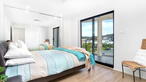A modern bedroom with a large bed, a view of a cityscape through a sliding door, and a wicker chair by the window. - Dolphin Cove Apartments, 13/2-6 Nelson Bay - 300m stroll to the marina! (Nelson Bay)