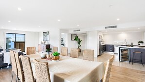 A modern dining area with a large table, wicker chairs, and a view of the kitchen. - Dolphin Cove Apartments, 13/2-6 Nelson Bay - 300m stroll to the marina! (Nelson Bay)