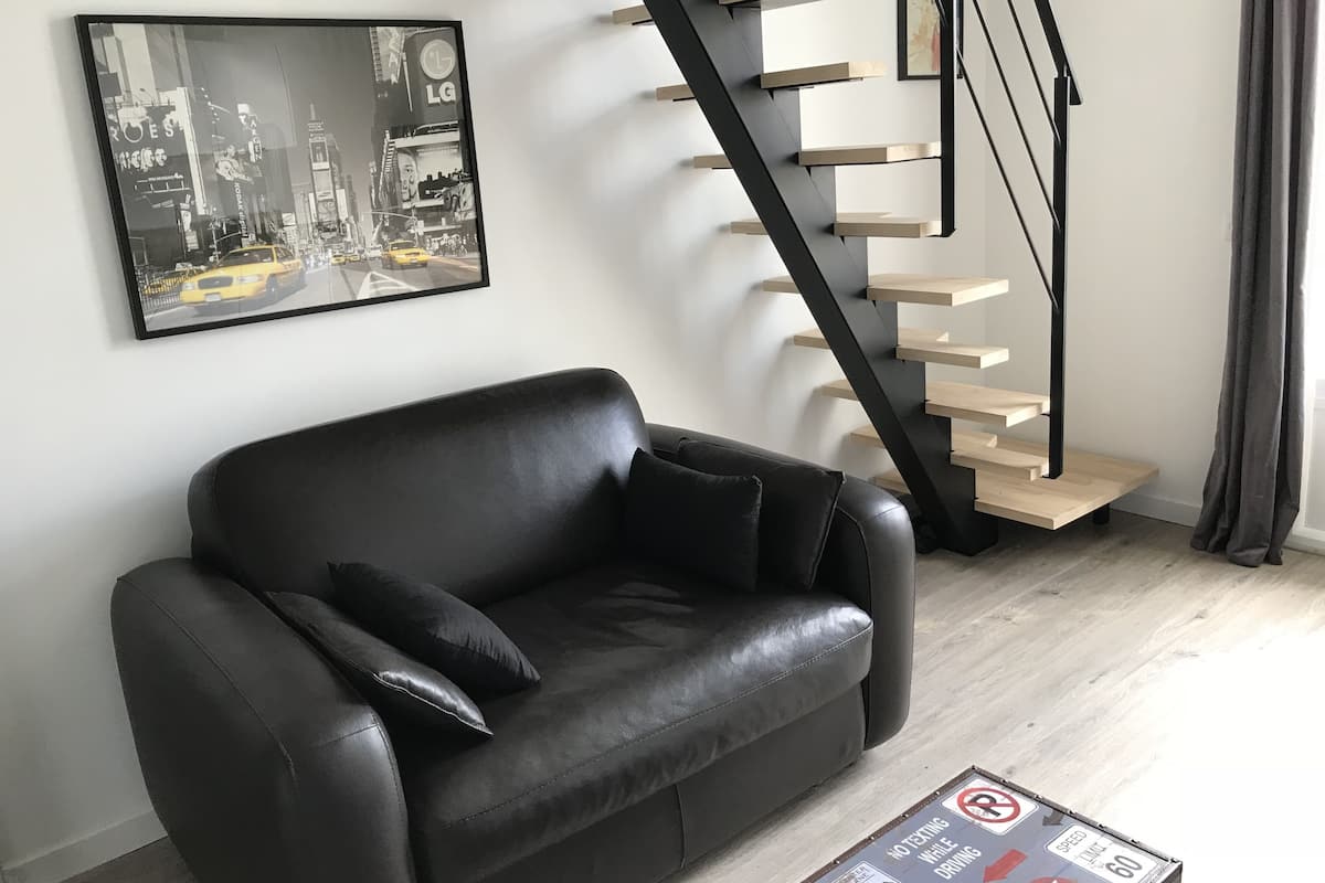 A modern living room with a black leather sofa, a wooden staircase, and a cityscape painting on the wall.