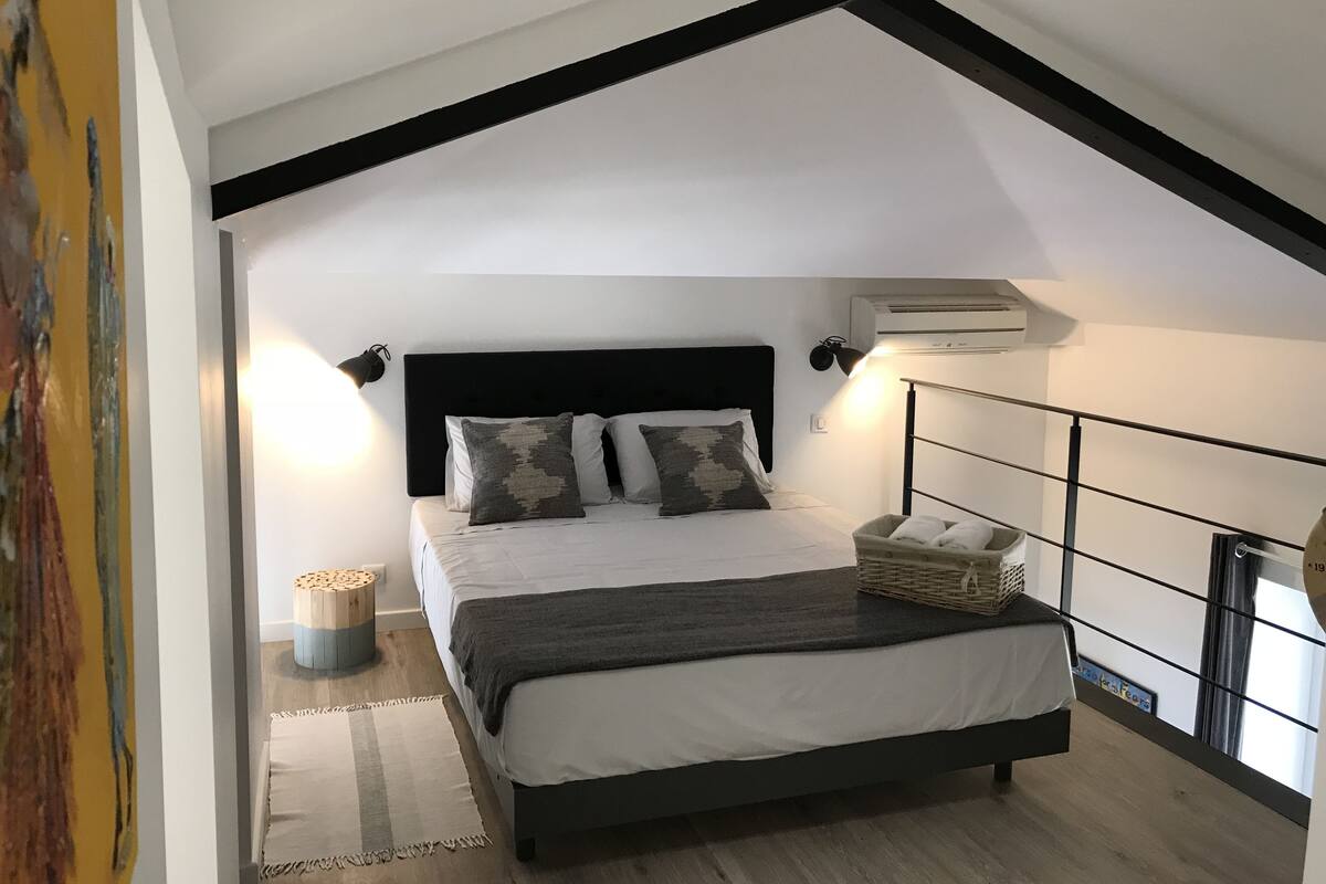 A bedroom with a bed, bedside lamps, a wall-mounted air conditioner, and a small side table with a basket.