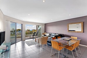 A modern living room with a glass dining table, leather sofa, and a balcony with a view. - Surfside 5 Fingal Bay Apartment with Water Views (Fingal Bay)