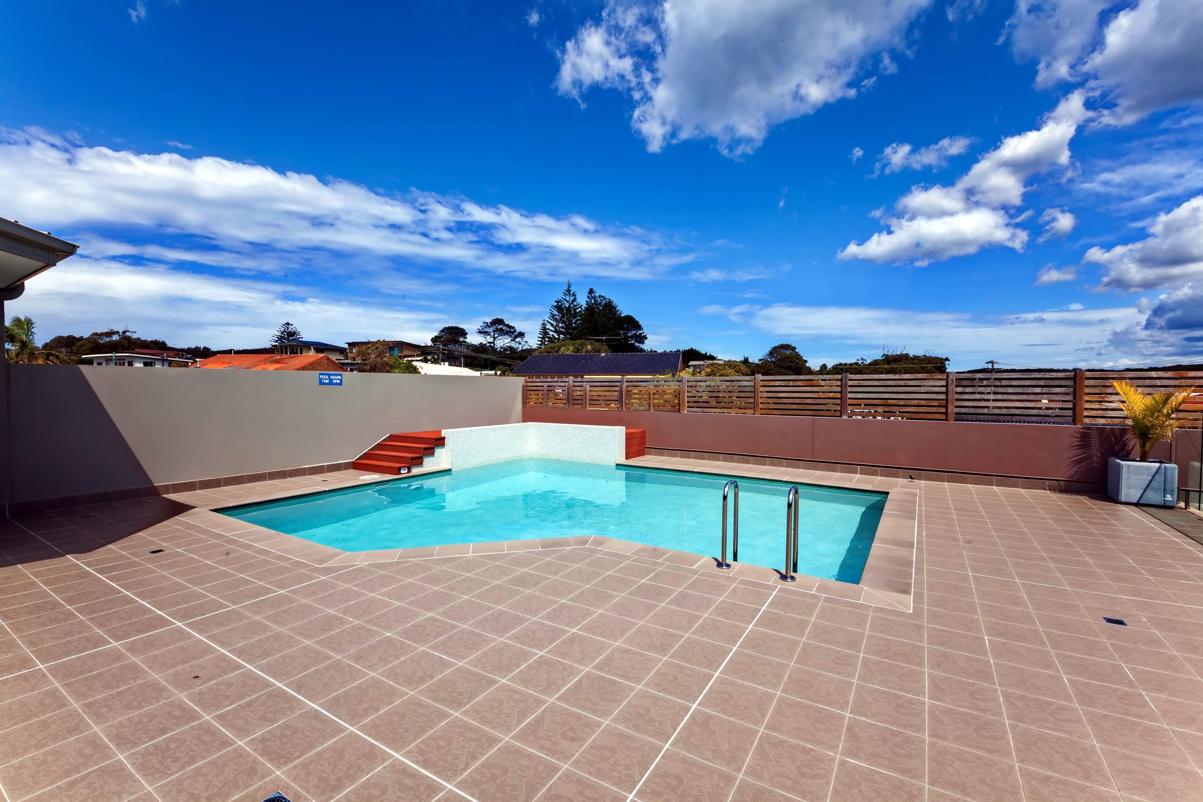Une piscine rectangulaire, bordée d’une terrasse carrelée, entourée d’un petit muret et d’une clôture en bois.