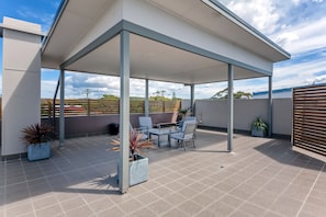 Een overdekt terras buiten, met betegelde vloer, twee stoelen en potten met planten.