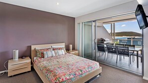 Quarto com cama grande, mesa de cabeceira, abajur e porta de vidro deslizante para uma varanda com mesa e cadeiras.
