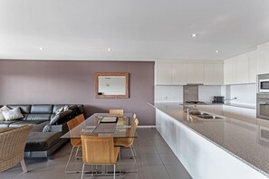 A modern living room with a glass dining table, leather sofa, and a kitchen with white cabinets and a stainless steel sink.