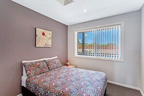 A bedroom with a bed, a window with blinds, a wall-mounted artwork, and a nightstand with a lamp. - Surfside 5 Fingal Bay Apartment with Water Views (Fingal Bay)