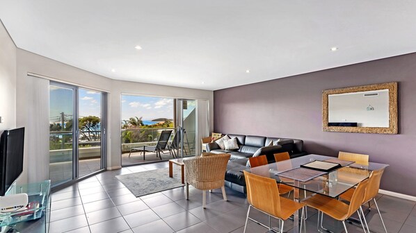A modern living room with a glass dining table, leather sofa, and a large window offering a view of the outdoors.