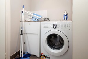 A compact laundry room with a front-loading washing machine, a mopping bucket, and a mop. - Surfside 5 Fingal Bay Apartment with Water Views (Fingal Bay)