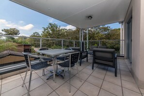 Een overdekt terras buiten met een tafel, stoelen, een barbecue en uitzicht op bomen en gebouwen.