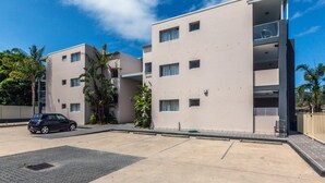 Um edifício residencial moderno de vários andares, com varandas, área de estacionamento e vegetação ao redor.